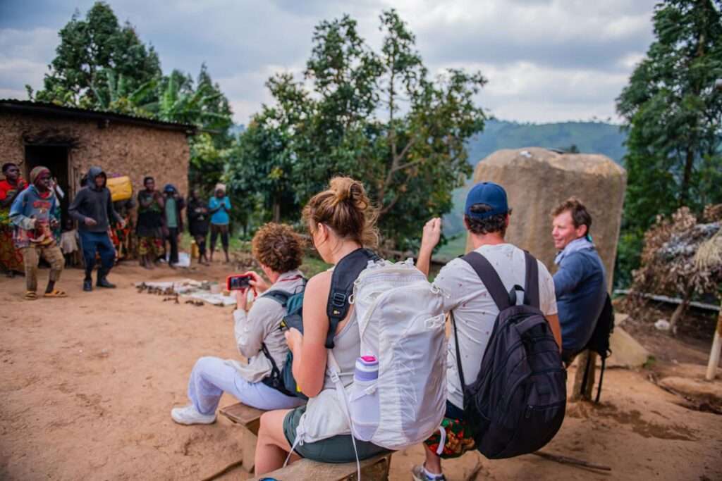 Several people with backpacks and cameras sit and observe a group of locals performing or gathering outside near trees and rustic buildings in a rural setting. The scene is lively, with some people watching and others participating.