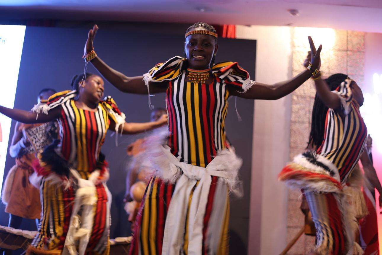 Women in brightly colored, striped traditional attire dance energetically with arms raised, performing on a stage with blurred background and soft lighting.