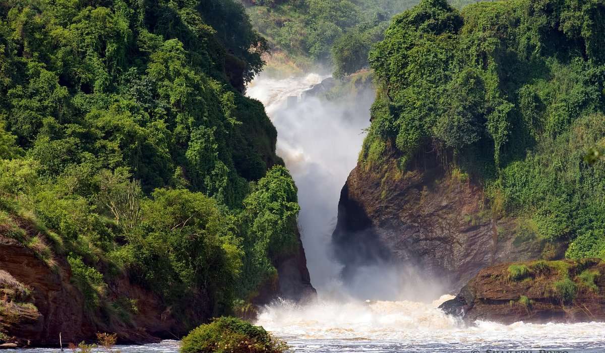 A powerful waterfall cascades between lush, green cliffs surrounded by dense forest, with mist rising from the crashing water below and sunlight filtering through the trees.